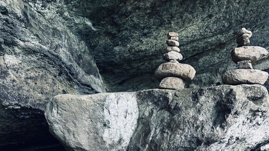 Two small towers of smooth stones are stacked on a large rock ledge beneath an overhanging rocky formation, creating a sense of balance and tranquility in a natural, rugged setting.